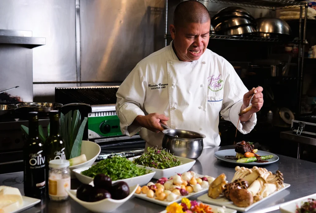 Chef plating a gourmet entrée in a commercial kitchen surrounded by fresh ingredients and prepared dishes.