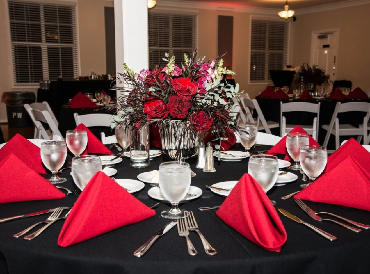 Elegant holiday event table setting with red napkins, black tablecloth, glassware, and a floral centerpiece of red and greenery in a warmly lit dining space.