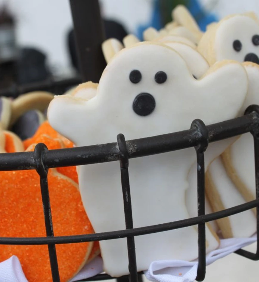 Close-up of Halloween-themed cookies shaped like white ghosts with black eyes and mouths, displayed in a basket alongside orange sugar-coated treats.