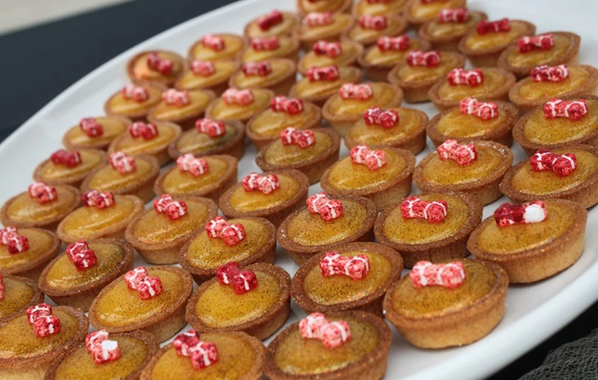 Tray of bite-sized dessert tarts topped with small red candy pieces, arranged neatly for a catered event or holiday gathering.